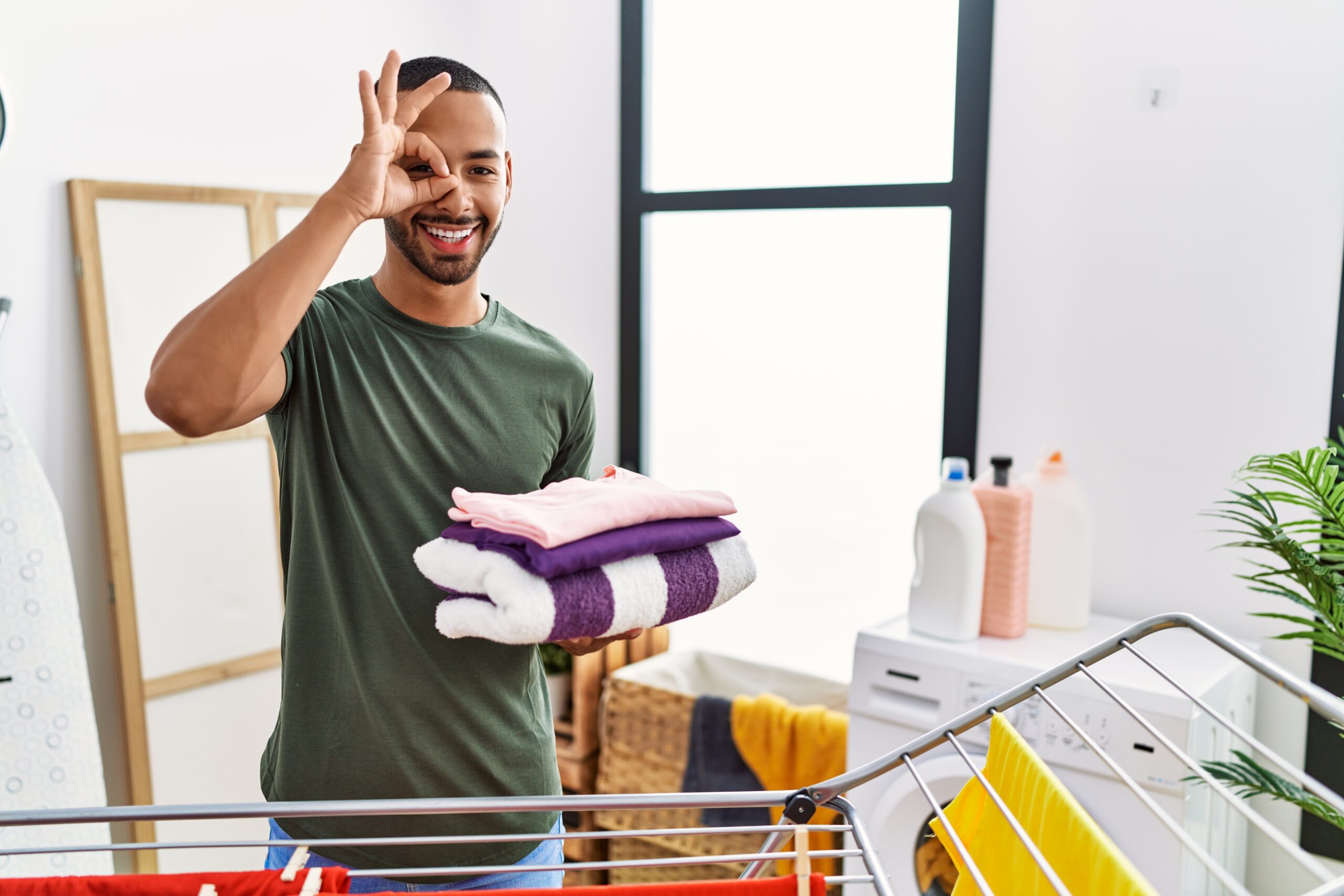 african american man holding folded laundry from clothline smiling happy doing ok sign with hand eye looking through fingers scaled